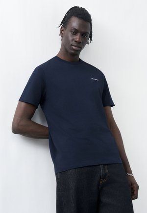 Un jeune homme aux cheveux tressés s'appuie contre un mur blanc, portant un t-shirt Calvin Klein bleu marine et un jean en denim foncé.