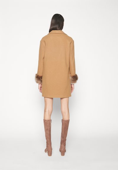 Cappotto color cammello con polsini in pelliccia, caratterizzato da una vestibilità dritta e colletto a rever. Indossato con stivali in suede fino al ginocchio.