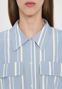 Camicia azzurro chiaro con strisce verticali bianche e nere, colletto a punta e due tasche sul petto con chiusura a bottone.