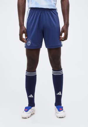 Mann trägt blaue Sportshorts, marineblaue knielange Socken mit weißen Streifen und weiße Fußballschuhe mit blauen und roten Akzenten.