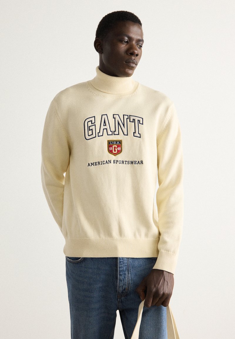 Cremfarbener Rollkragenpullover aus weichem Material, mit blauer gestickter "GANT"-Logoschrift und dem Text "AMERICAN SPORTSWEAR". Getragen mit blauen Jeans.