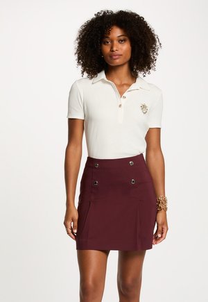 Donna con capelli ricci che indossa una polo bianca a maniche corte e una gonna bordeaux a vita alta con dettagli dorati sui bottoni, in piedi su sfondo bianco.
