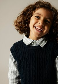 Suéter de punto azul marino con un patrón de trenzas, superpuesto a una camisa de cuello a rayas en azul claro y blanco. El niño tiene el cabello rizado y dientes visibles.