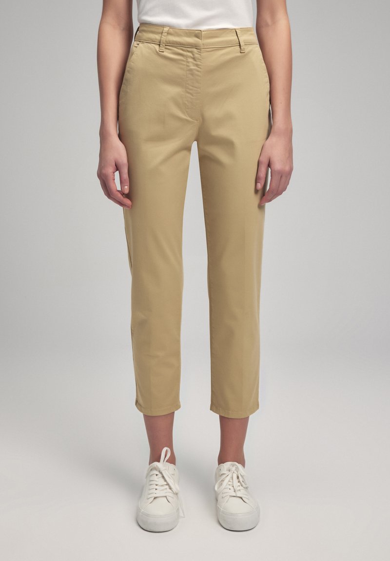 Pantaloni cropped beige con una texture liscia, vestibilità slim e tasche laterali, abbinati a sneakers bianche per un look casual.