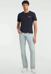 Junger Mann in einem schwarzen T-Shirt mit kurzen Ärmeln, hellblauen Jeans, braunem Gürtel und weißen Sneakern, der mit einer Hand in der Tasche steht.