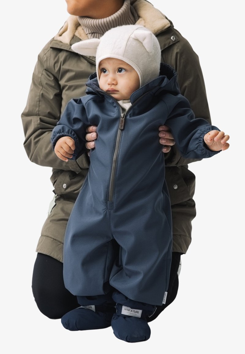 Dunkelblauer Kinder-Schneeanzug mit Reißverschluss, elastischen Bündchen und angenähten Fußteilen, aus wasserdichtem Material. Weiche weiße Mütze mit Ohrenakzenten.
