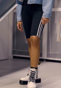 Shorts de cyclisme noirs avec des rayures blanches, associés à des bottines blanches à lacets avec une épaisse semelle noire et une languette jaune.