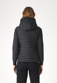 Persona con cabello castaño de longitud media, vistiendo una chaqueta con capucha acolchada negra y pantalones negros, vista desde atrás contra un fondo blanco.