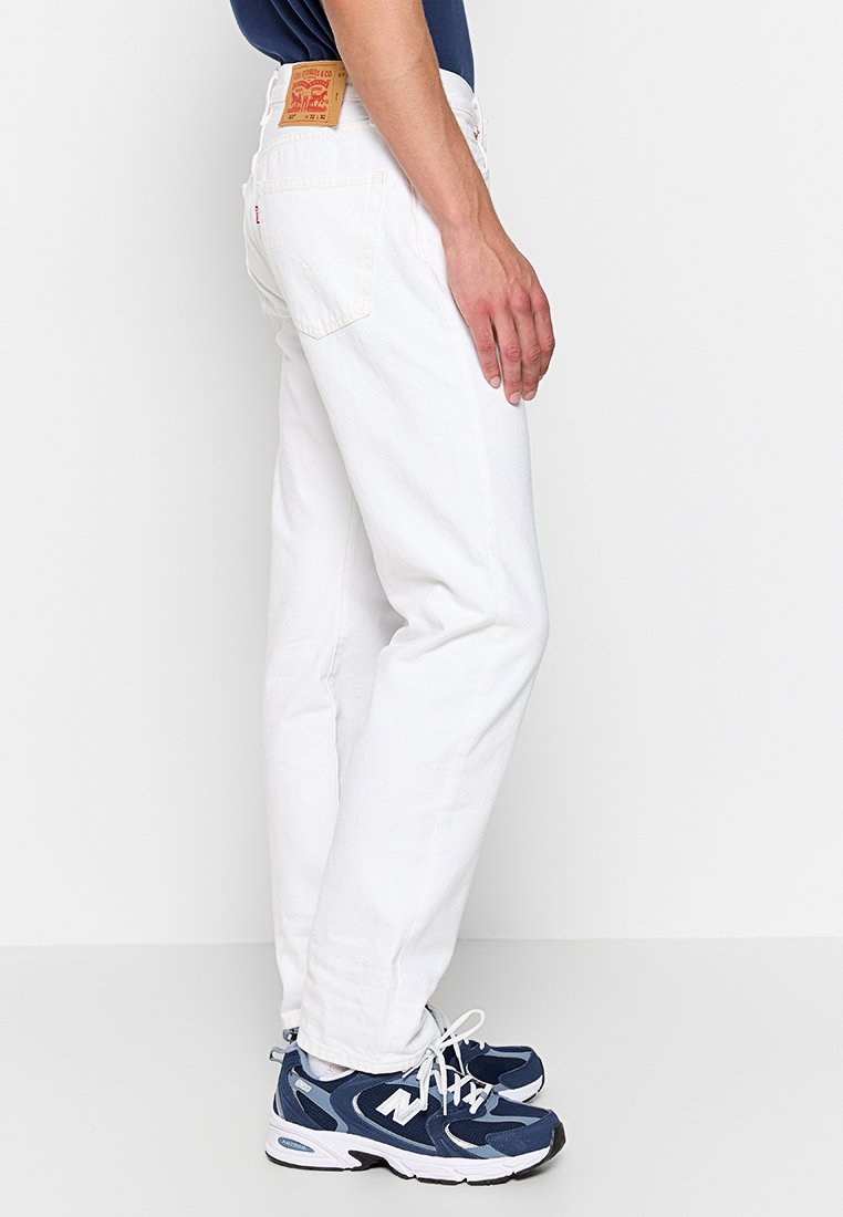 Weiße Denim-Jeans mit geradem Schnitt, strukturiertem Stoff und einem Leder-Logo-Patch am hinteren Bund. Die Schuhe sind im dunkelblauen Sportstil.