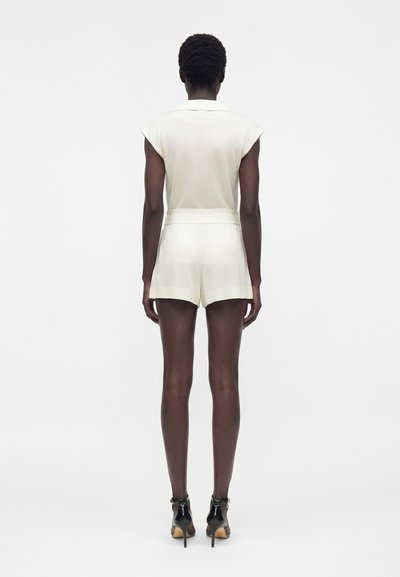 Γυναίκα που στέκεται με την πλάτη, φοράει ένα λευκό αμάνικο ολόσωμο με γιακά και ζώνη, σε συνδυασμό με μαύρα πέδιλα με ψηλό τακούνι.