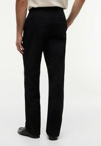 Uomo che indossa pantaloni neri a gamba dritta e scarpe eleganti nere, in piedi con una mano in tasca, sfondo bianco.