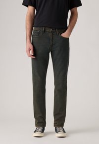 Hombre vestido con jeans ajustados desgastados negros, camiseta negra y zapatillas negras con cordones blancos, de pie con una mano en el bolsillo.