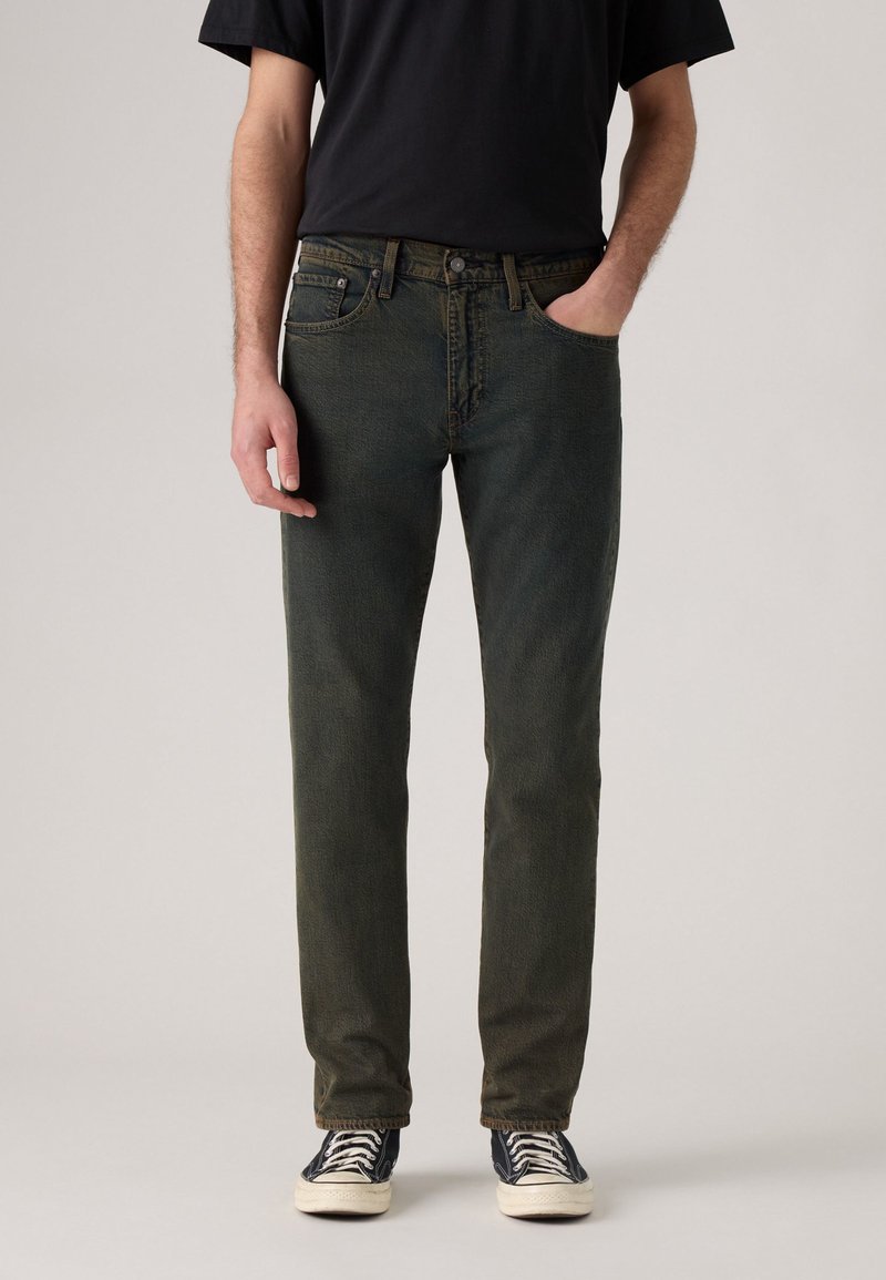 Hombre vestido con jeans ajustados desgastados negros, camiseta negra y zapatillas negras con cordones blancos, de pie con una mano en el bolsillo.