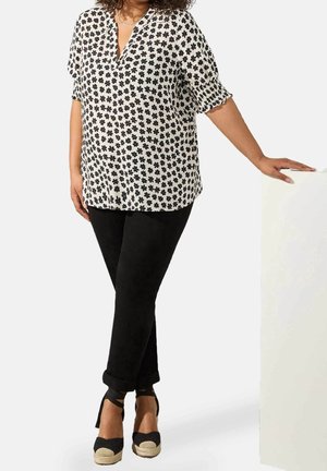 Vrouw draagt een witte blouse met zwarte bloemenprint, zwarte broek en zwarte sleehakken, staand en leunend op een wit oppervlak.
