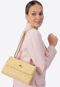 Żółta pikowana torebka na ramię z paskiem z złotego ła�ńcuszka i klapą zakładaną na przód. Na froncie znajduje się ozdoba z logo, a powierzchnia jest gładka.