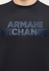 Κοντινή λήψη μπλε σκούρου πουκαμίσου με υφή, με μπλε και γκρι λογότυπο "ARMANI EXCHANGE" στο στήθος.