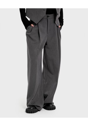 Osoba nosząca luźne szare plisowane spodnie, czarne masywne buty oraz szarą kamizelkę na czarnym topie z długim rękawem, ręce w kieszeniach.