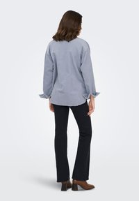 Naine seisab seljaga, kandes helehalli korduroost särki, mustad lehvitavate säärega teksapükse ja pruune kontsaga poolsaabast.