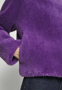 Lila fuskpäls tröja med en mjuk, strukturerad yta, raglanärmar och en kortare passform. Har sidofickor och en rundad fåll.