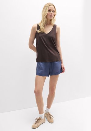 Frau steht drinnen und trägt ein dunkelbraunes ärmelloses V-Ausschnitt-Top, blaue Shorts, weiße Socken und beige Sneakers.