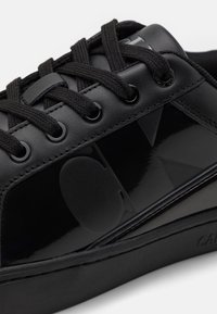 Zbliżenie czarnego buta sportowego z teksturowanymi sznurowadłami, błyszczącą boczną częścią i wytłoczonym okrągłym logo na boku.