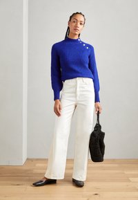 Bright blue ribbed knit sweater with a high neckline and button detail, paired with wide-leg white trousers and a textured black bag.