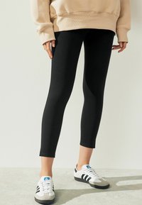 Zwarte leggings met een aansluitend ontwerp, gemaakt van een rekbaar materiaal, gecombineerd met een beige oversized sweatshirt en witte sportschoenen met grijze accenten.