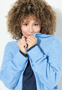 Ljusblå stickad cardigan med ribbade muddar; mjuk textur; bärs stängd runt halsen; marinblå topp under; lockigt hår som ramar in ansiktet.
