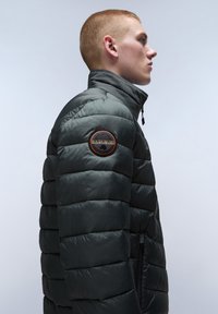 Puffjacka i mörkgrön, med quiltat mönster, hög krage och en svart cirkelmärke på axeln. Dragkedjedetalj synlig.