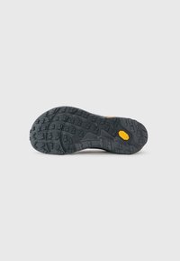 Schwarze Gummisohle eines Schuhs mit tiefen Profilen und einem gelben Vibram-Logo near der Ferse auf einem schlichten weißen Hintergrund.