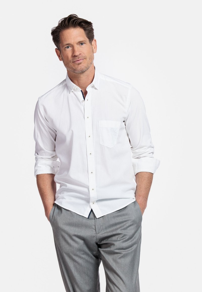 Witte knoopshirt met een enkele borstzak, opgerolde mouwen en grijze op maat gemaakte broek, weergegeven tegen een effen achtergrond.