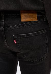 Κοντινό πλάνο μαύρου τζιν Levi's Lot 502 που δείχνει την πίσω τσέπη, την κόκκινη ετικέτα Levi's και το καφέ δερμάτινο σήμα με το μέγεθος W31 L34.