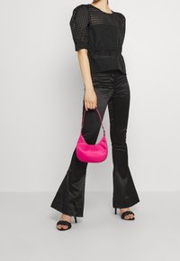 Top nera a maniche a sbuffo con vita arricciata, abbinato a pantaloni neri lucidi a zampa d'elefante. Tiene in mano una piccola borsa in raso rosa brillante.