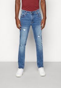 Blue denim jeans with a slim fit, featuring distressed details and a standard five-pocket design, paired with white sneakers.