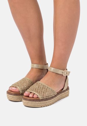 Pies con sandalias de plataforma beige de puntera abierta, con textura tejida y correas con hebillas en el tobillo, sobre un fondo liso claro.