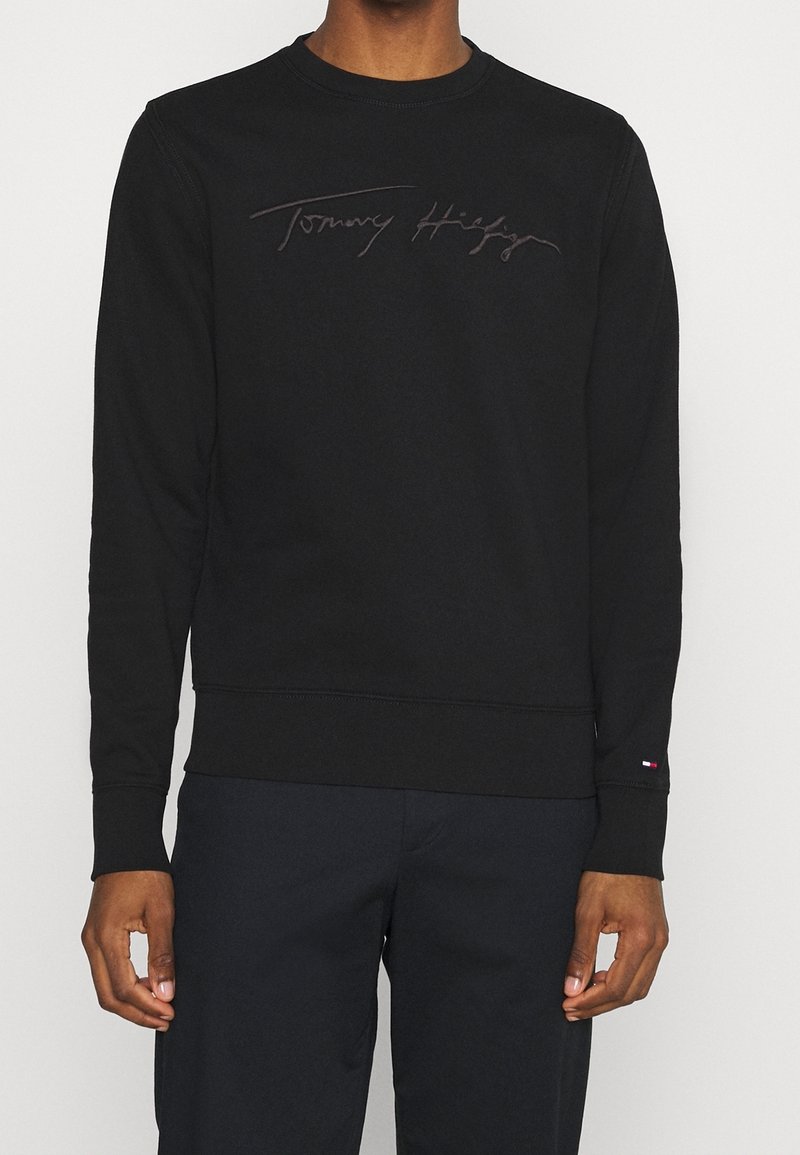 Personne portant un sweat-shirt à manches longues noir avec le texte "Tommy Hilfiger" sur la poitrine et un pantalon foncé sur un fond uni.