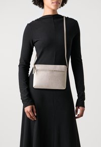 Světlé béžové kožené crossbody taška s texturovaným povrchem, obdélníkovým tvarem, zipovým uzavřením a štíhlým nastavitelným popruhem.