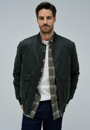 Un homme portant une veste bomber vert foncé par-dessus une chemise à carreaux verte et un tee-shirt blanc, se tenant contre un fond gris clair uni.