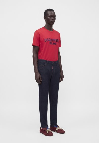 Άνδρας που στέκεται, φοράει κόκκινο T-shirt Dsquared2 Milano, σκούρο μπλε τζιν και κόκκινα αθλητικά με μπεζ κορδόνια σε λευκό φόντο.