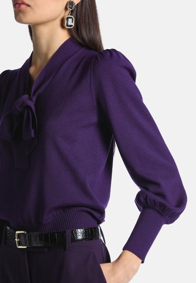 Femme portant un chemisier violet à manches longues avec un nœud au col, des manches bouffantes et une ceinture noire texturée sur un pantalon foncé.