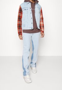 Light blue denim vest with red and yellow plaid sleeves over a brown hoodie, paired with light blue jeans featuring side panels and white sneakers.
