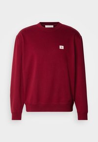 Sweatshirt bordeaux à col rond avec manches longues et petit patch logo "CK" blanc et rouge sur le côté gauche de la poitrine.