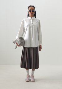 Cămașă albă cu guler și mâneci lungi, fustă plisată maro, adidași roz deschis și geantă gri. Ochelarii de soare negri completează ținuta.