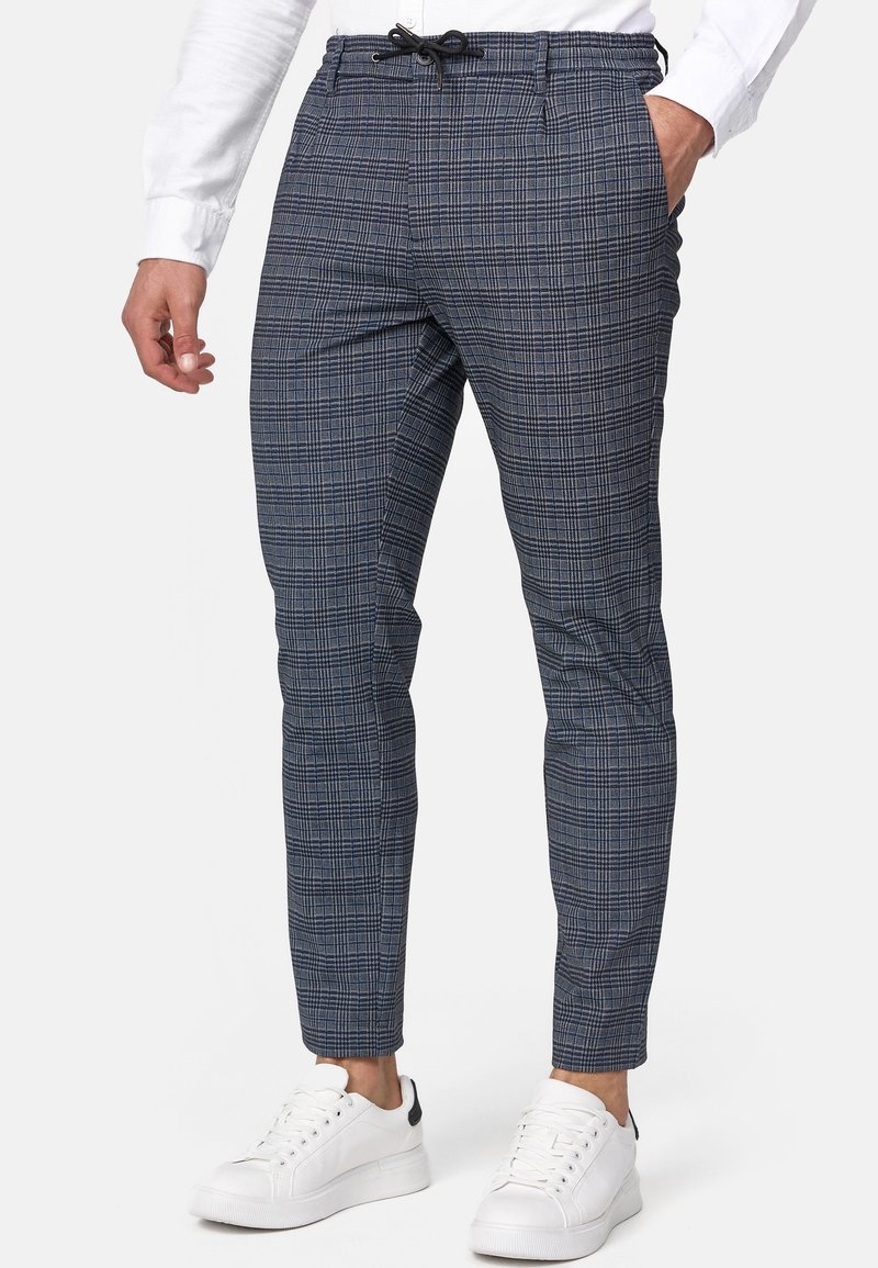 Pantalon slim gris à motif à carreaux avec cordon noir porté par une personne en baskets blanches et chemise blanche à manches longues.