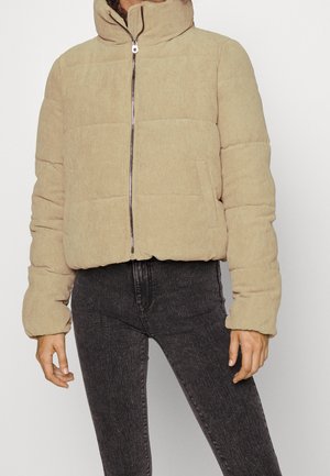 Person trägt eine beige Cord-Steppjacke mit hohem Kragen und schwarz gewaschene Skinny-Jeans, steht vor einem einfarbigen Hintergrund.