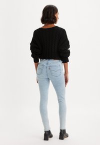 Egy nő áll, háttal a kamerának, fekete, kötött pulóverben, világoskék skinny farmernadrágban és fekete bokacsizmában, fehér háttér előtt.