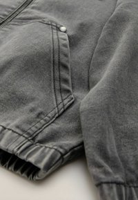 Chaqueta de mezclilla gris con cierre de cremallera, botones a presión, puños elásticos y tejido texturizado con detalles de costura visibles.