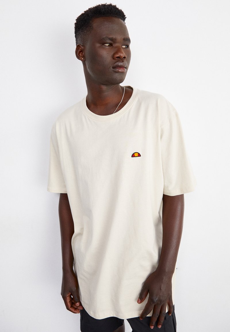Helles beiges Baumwoll-T-Shirt mit rundem Ausschnitt, kurzen Ärmeln und kleinem, mehrfarbigen Logo auf der Brust. Glatte Textur.