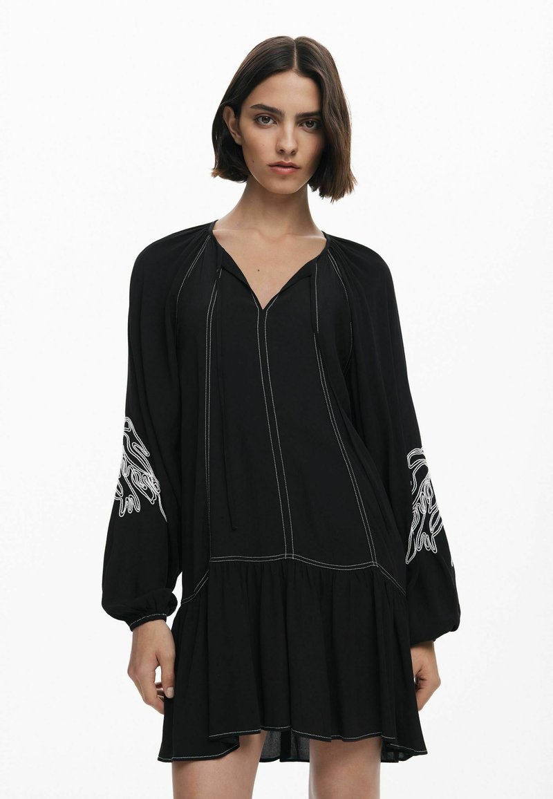 Schwarzes Kleid mit langen Ärmeln, verziert mit weißen gestickten Akzenten und einem ausladenden Saum. Hergestellt aus leichtem Stoff mit einem V-Ausschnitt.