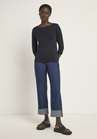 Schwarzes 3/4-Ärmel-Oberteil kombiniert mit blauen weit geschnittenen Jeans mit Denim-Bündchen. Die Schuhe sind schwarze Slidesandaletten. Einfaches, lässiges Outfit.